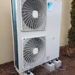 Zamontowana zewnętrzna jednostka pompy ciepła Daikin Altherma na betonowych podstawach z kółkami, ustawiona przy beżowej ścianie budynku i iglaku.