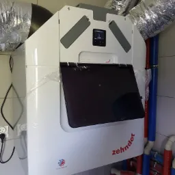 Biała jednostka rekuperacyjna Zehnder z podłączonymi przewodami wentylacyjnymi i instalacją hydrauliczną w pomieszczeniu z kafelkami, jeszcze w folii ochronnej.