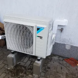 Biała jednostka zewnętrzna klimatyzatora Daikin, zamontowana na betonowych bloczkach, z widocznymi rurami odprowadzającymi skropliny i resztkami wody na obudowie, ustawiona na kostce brukowej przy...