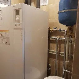 Biała jednostka grzewcza Atlantic Alfea Extensa z niebieskim zbiornikiem wyrównawczym i rurami w pomieszczeniu wyłożonym kafelkami.