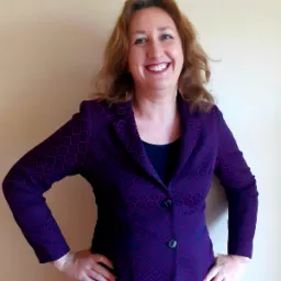 Uśmiechnięta kobieta w bordowo-fioletowej marynarce z geometrycznym wzorem, opierająca dłonie na biodrach na tle beżowej ściany.