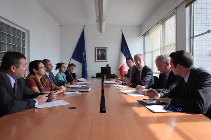 Grupa osób w garniturach siedzi przy długim, drewnianym stole w sali konferencyjnej z flagami Unii Europejskiej i Francji w tle, sugerując spotkanie biznesowe lub negocjacje.