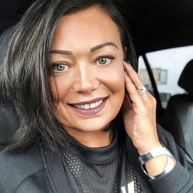 Uśmiechnięta brunetka z pierścionkiem V na palcu, w czarnej bluzie, siedzi w samochodzie. Subtelny makijaż i srebrna bransoletka dodają blasku.