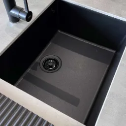 Czarny, kwadratowy zlew kuchenny z ociekaczem i czarną baterią, zamontowany w blacie o jasnoszarej teksturze betonu.