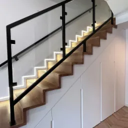 Drewniane schody z podświetleniem LED i szklaną balustradą z czarnymi elementami, zabudowane szafy wnękowe pod schodami, parkiet ułożony w jodełkę.