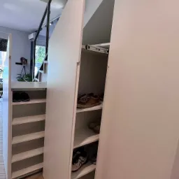 Biała szafa na buty na wymiar, z wysuwanymi półkami, umieszczona pod schodami, widoczne buty na półkach, parkiet na podłodze.