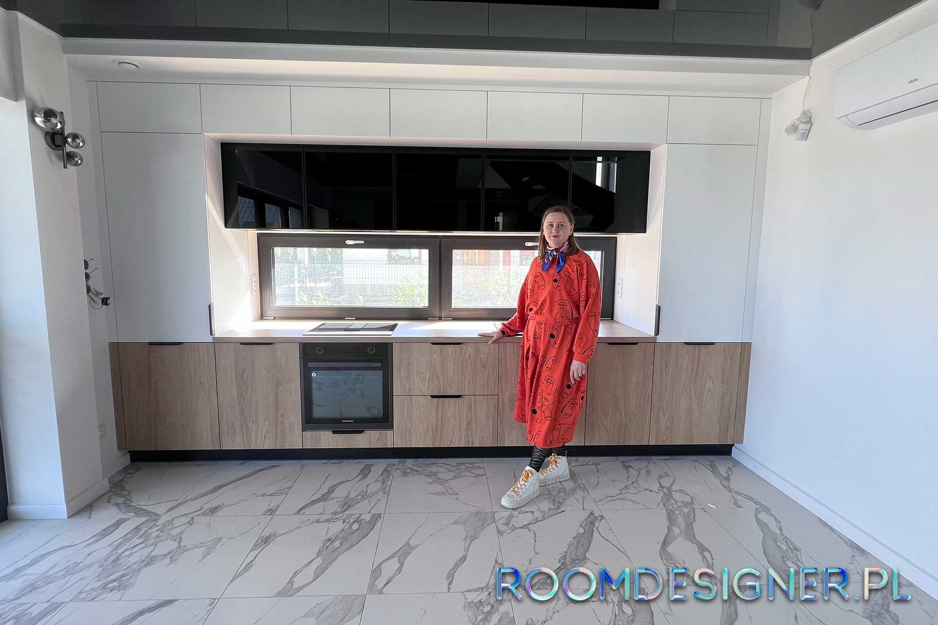 Jasna kuchnia z drewnianymi frontami szafek, czarnymi elementami i marmurową podłogą; kobieta w czerwonej sukience stoi na tle zabudowy.