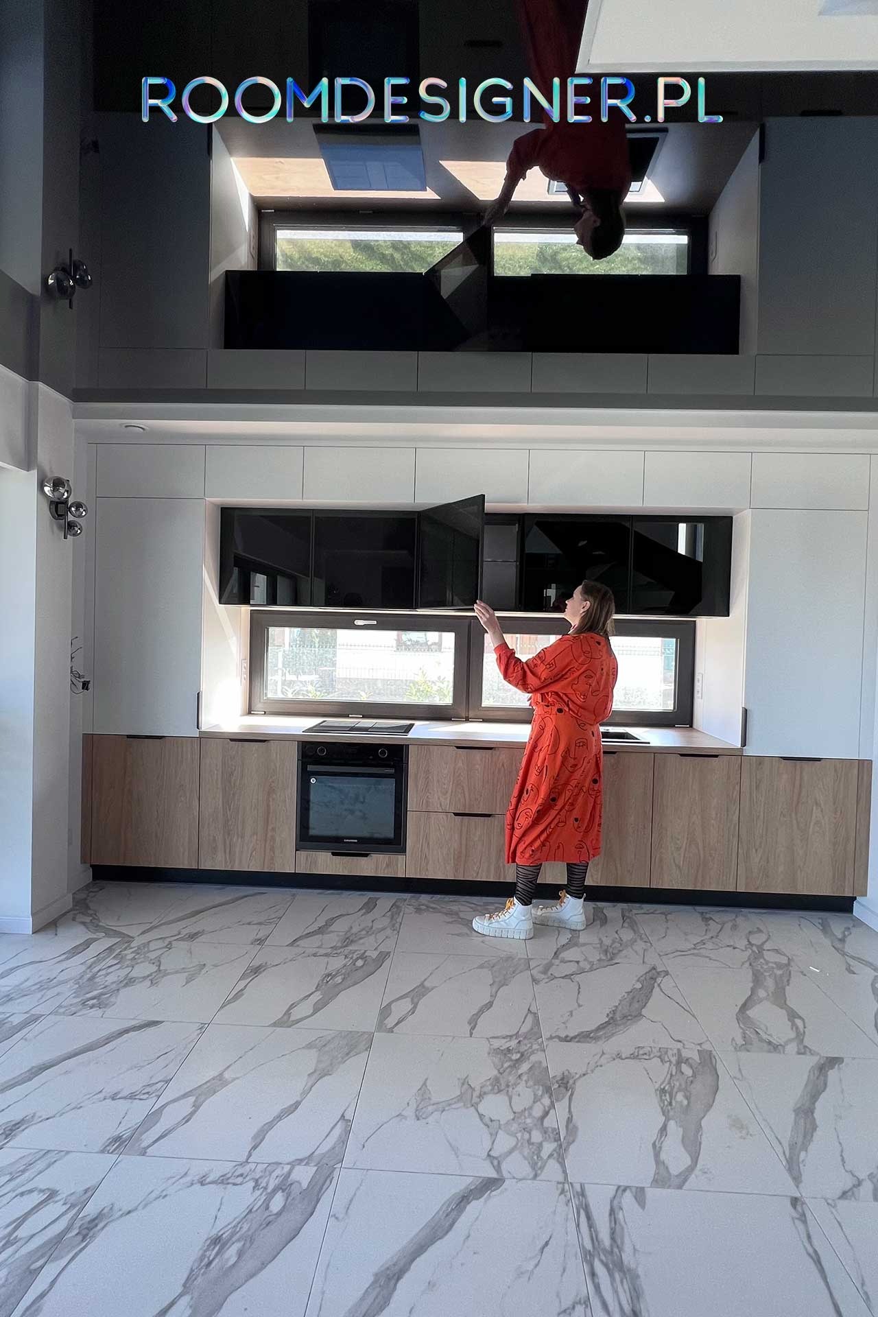 Kobieta w pomarańczowej sukience otwiera szafkę w nowoczesnej kuchni z białymi górnymi szafkami i drewnopodobnymi dolnymi szafkami, marmurowa podłoga, nad szafkami logo firmy projektującej wnętrza.