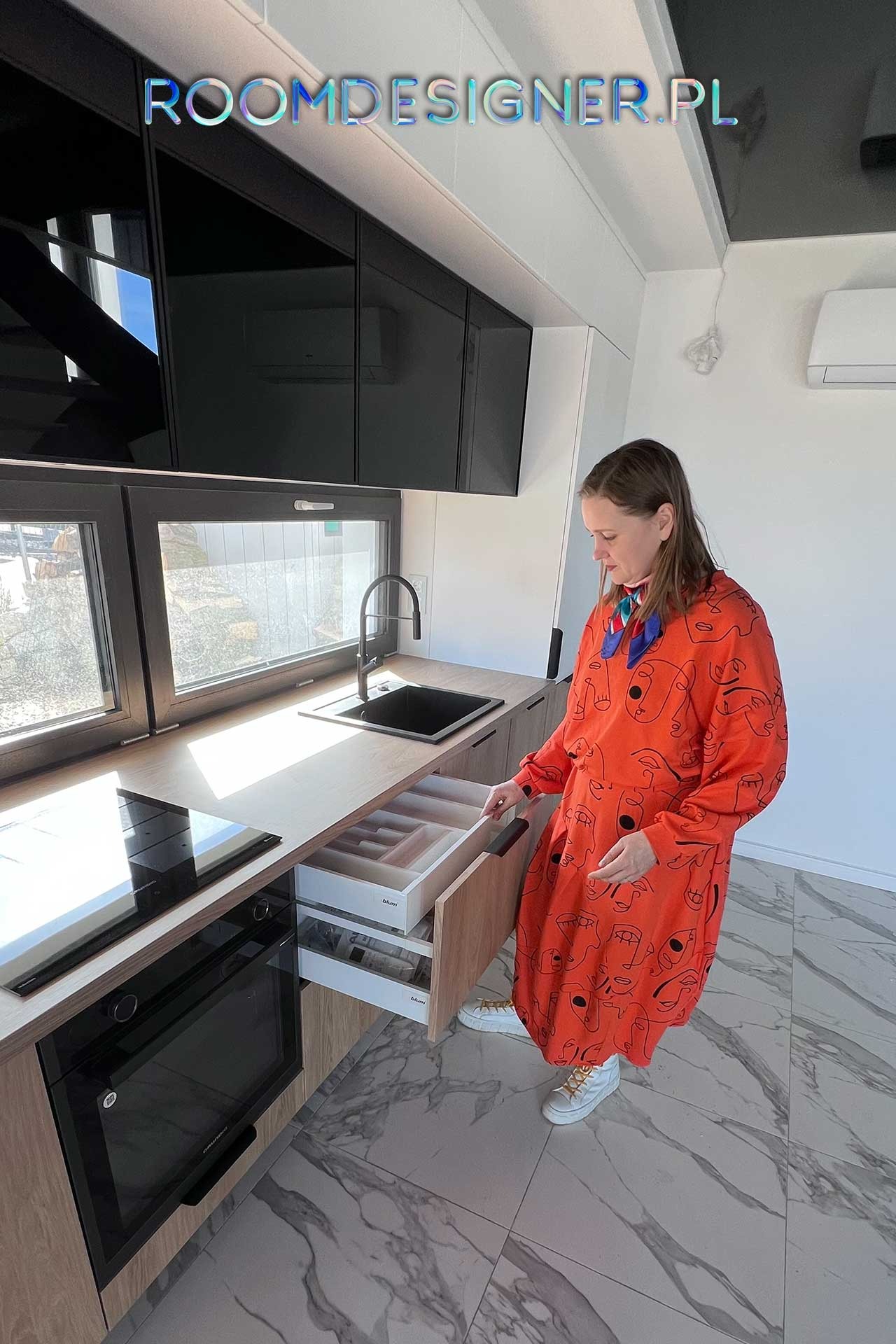 Kobieta w pomarańczowej sukience otwiera szufladę z przegródkami na sztućce w nowoczesnej kuchni z drewnianymi blatami i czarnymi szafkami wiszącymi.