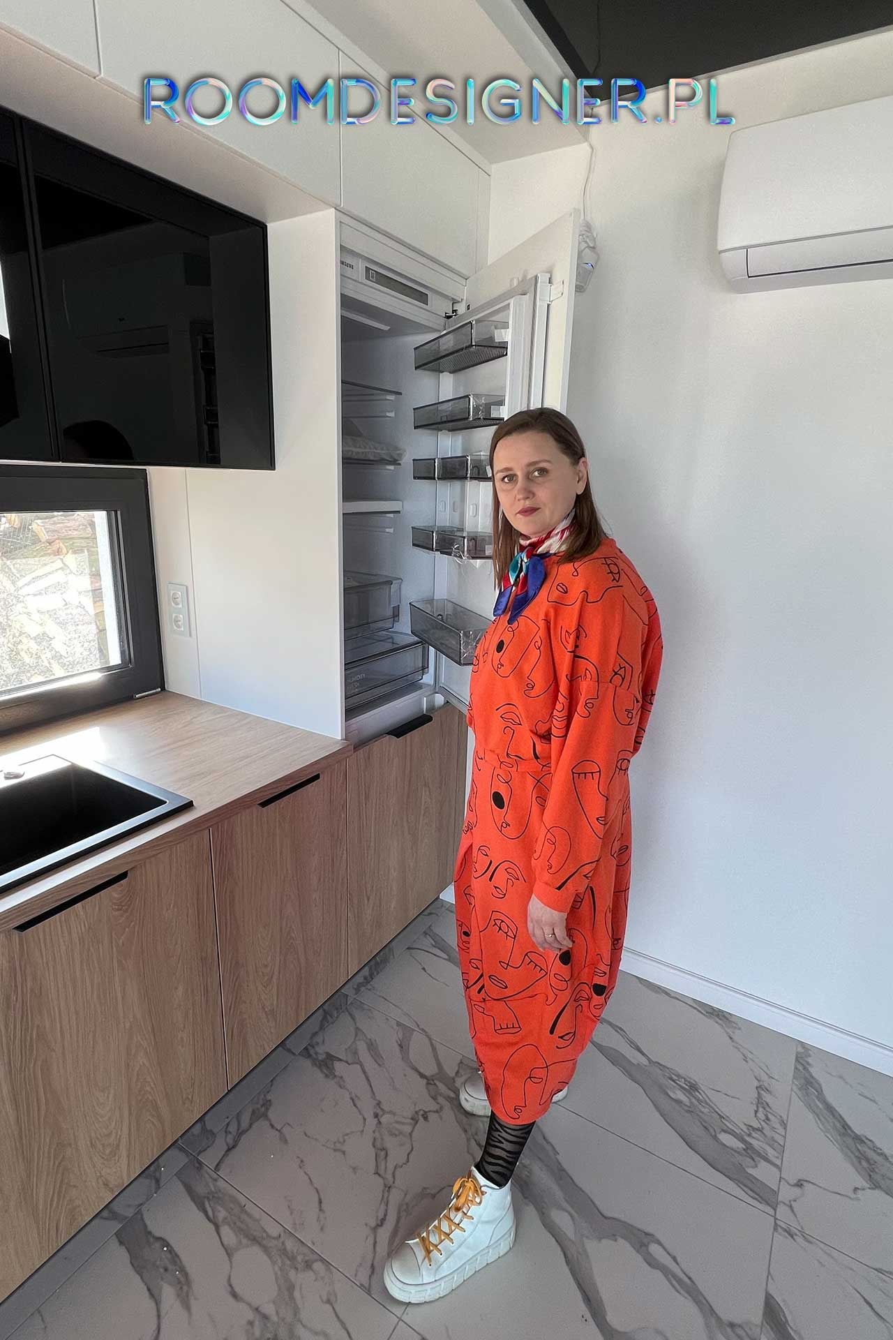 Kobieta w pomarańczowej sukience stoi w nowoczesnej kuchni z drewnianymi szafkami i otwartą lodówką, na podłodze marmurowe płytki.
