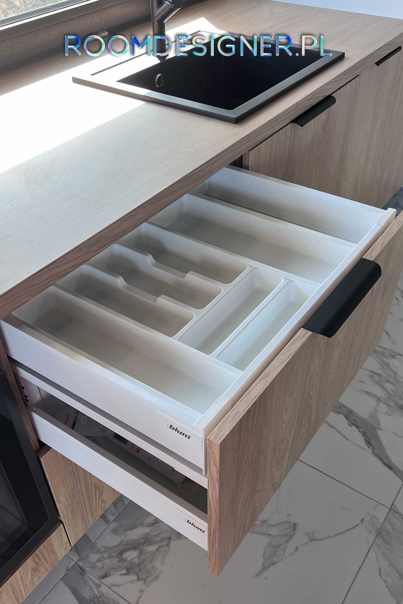 Otwarta szuflada kuchenna z wkładem na sztućce, fronty meblowe z drewnopodobnym wzorem, widoczny zlew i bateria w tle.