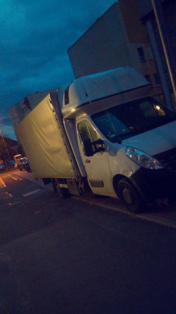 Biały samochód dostawczy z plandeką zaparkowany na ulicy o zmierzchu, widoczny budynek w tle.