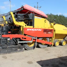 Żółto-czerwona rozkładarka asfaltu Dynapac F181-8W na placu budowy, widoczne elementy robocze i opony, w tle drzewa i składowisko materiałów.