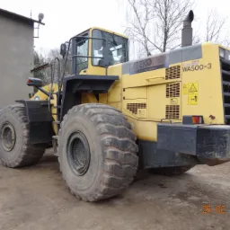 Żółta ładowarka kołowa Komatsu WA500-3 na placu budowy, widoczne duże opony, kabina operatora i łyżka. Na nadwoziu naklejka z logo firmy Abeko. W tle budynek gospodarczy i drzewa bez liści.