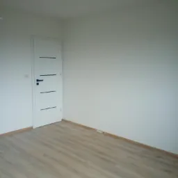 Pusty pokój po remoncie z panelami podłogowymi w odcieniach brązu i białymi ścianami. Białe drzwi z czarnymi poziomymi zdobieniami i klamką. Listwy przypodłogowe w kolorze drewna.