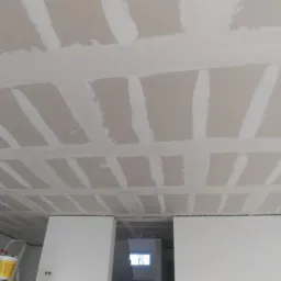 Sufit z płyt kartonowo-gipsowych w trakcie montażu, widoczne łączenia wypełnione masą szpachlową, fragment drabiny malarskiej w rogu pomieszczenia.