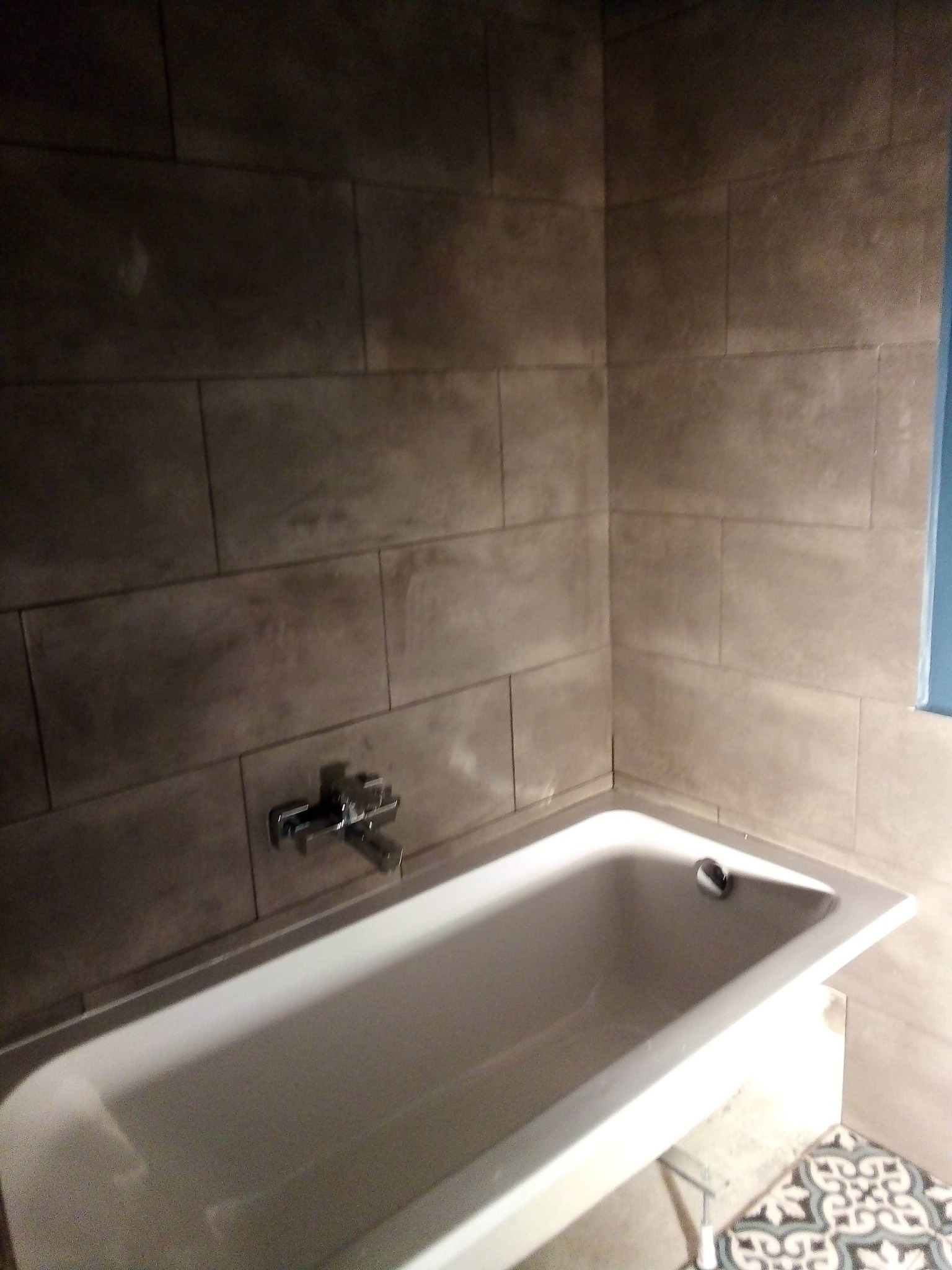 Wykończona łazienka z wanną obłożoną szarymi, prostokątnymi płytkami, bateria zamontowana na ścianie, widoczny fragment podłogi z wzorzystymi kaflami.