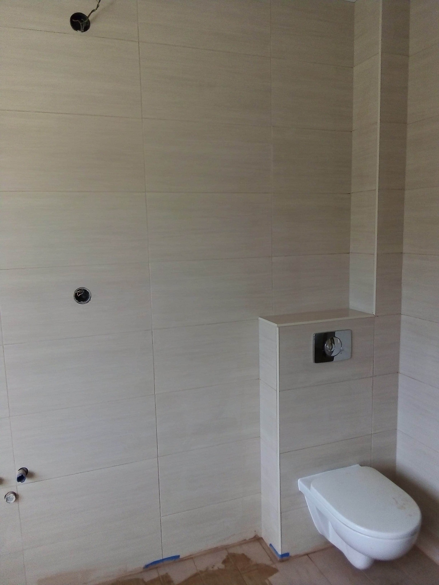 Ściana łazienki wyłożona beżowymi płytkami, widoczna podtynkowa spłuczka z przyciskiem i wisząca toaleta. Na podłodze widoczne ślady po pracach wykończeniowych.