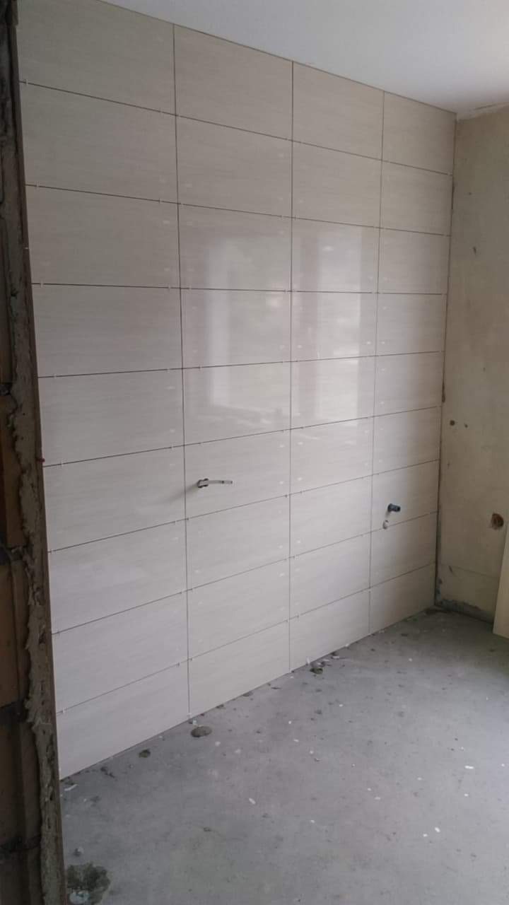 Ściana wyłożona jasnymi, prostokątnymi płytkami ceramicznymi z widocznymi otworami na instalacje, surowy beton na podłodze, fragment ościeżnicy z resztkami zaprawy.