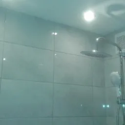 Wykończona łazienka z jasnymi, kwadratowymi płytkami, chromowaną deszczownicą i wentylatorem w rogu.