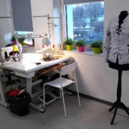 Wnętrze pracowni krawieckiej z maszyną do szycia pod lampą, krzesłem, manekinem z marynarką w geometryczny wzór i doniczkami z roślinami na parapecie okiennym.