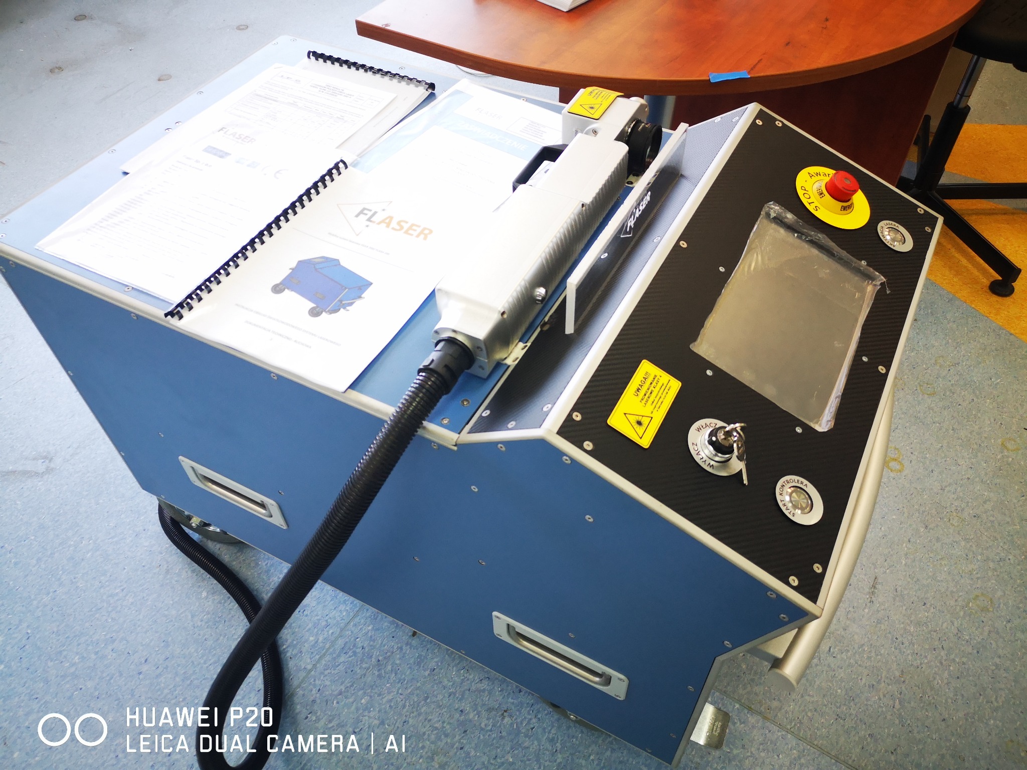 Przenośne urządzenie przemysłowe z głowicą laserową, panelem sterowania z przyciskiem awaryjnym i dokumentacją techniczną na górze, widoczne ostrzeżenia laserowe.