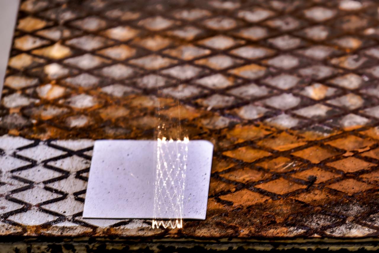 Metalowa kratownica z rdzawym nalotem, fragment białej kartki leżącej na powierzchni, odbijająca się w niej siatka, zbliżenie.