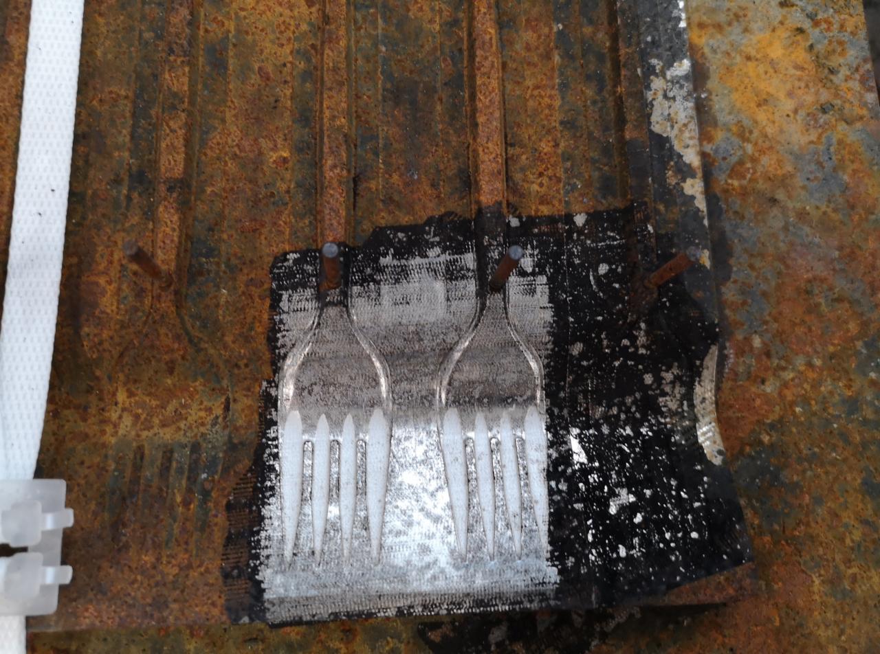 Metalowa powierzchnia z rdzawym nalotem, częściowo oczyszczona z użyciem szablonów w kształcie dwóch widelców, fragmenty czarnej farby i ślady po szlifowaniu.