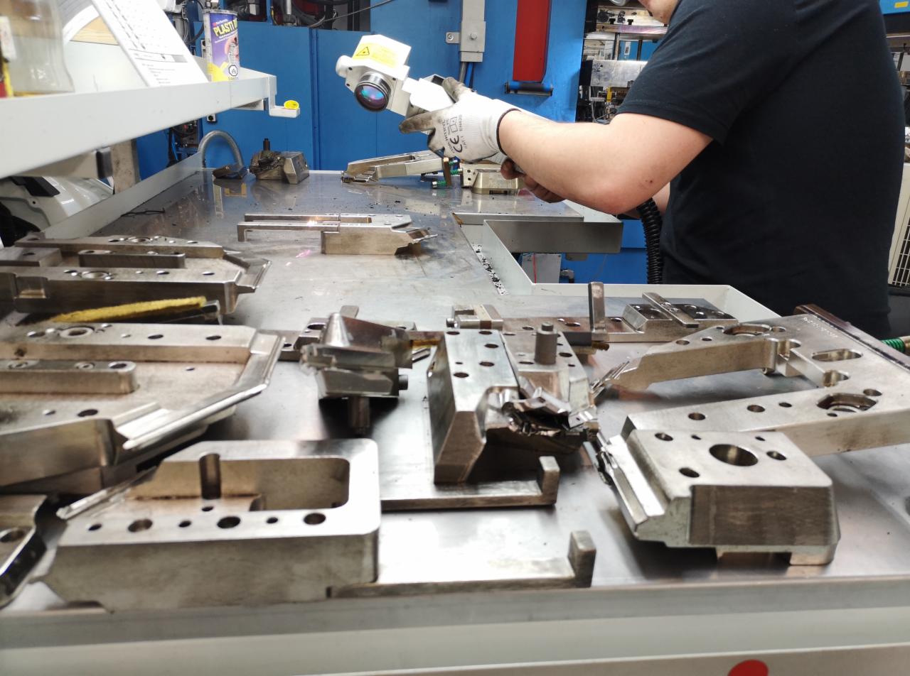 Metalowe elementy form obrabiane laserowo na stole warsztatowym, operator w rękawicy ochronnej używa urządzenia laserowego, widoczne ślady obróbki i drobne wióry.