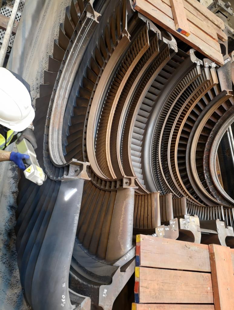 Przemysłowa turbina parowa w sekcji, skanowana przez pracownika w kasku i rękawicach ochronnych, widoczne drewniane podpory i szczegóły konstrukcyjne.