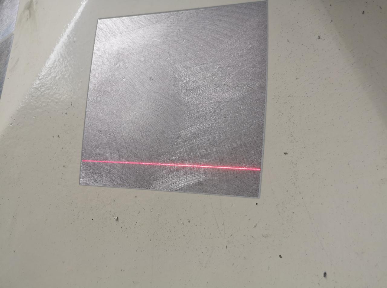 Metalowa płyta z widocznymi śladami obróbki i czerwoną linią lasera, prawdopodobnie w trakcie pomiarów lub znakowania.