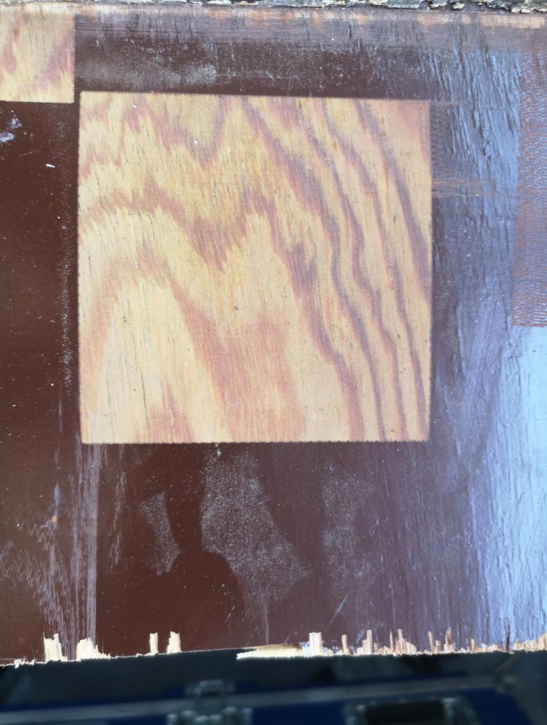 Fragment drewnianej powierzchni z kwadratowym obszarem po usunięciu starej farby, ukazującym naturalny rysunek drewna, otoczony brązową, częściowo uszkodzoną powłoką.