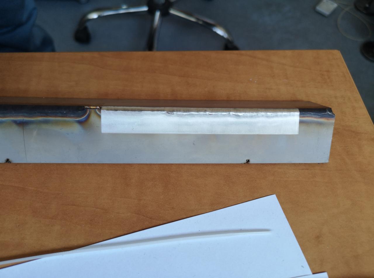 Metalowy profil z widocznymi śladami obróbki, częściowo zamaskowany taśmą przed dalszą obróbką, leżący na drewnianym stole, w tle widoczne krzesło biurowe.