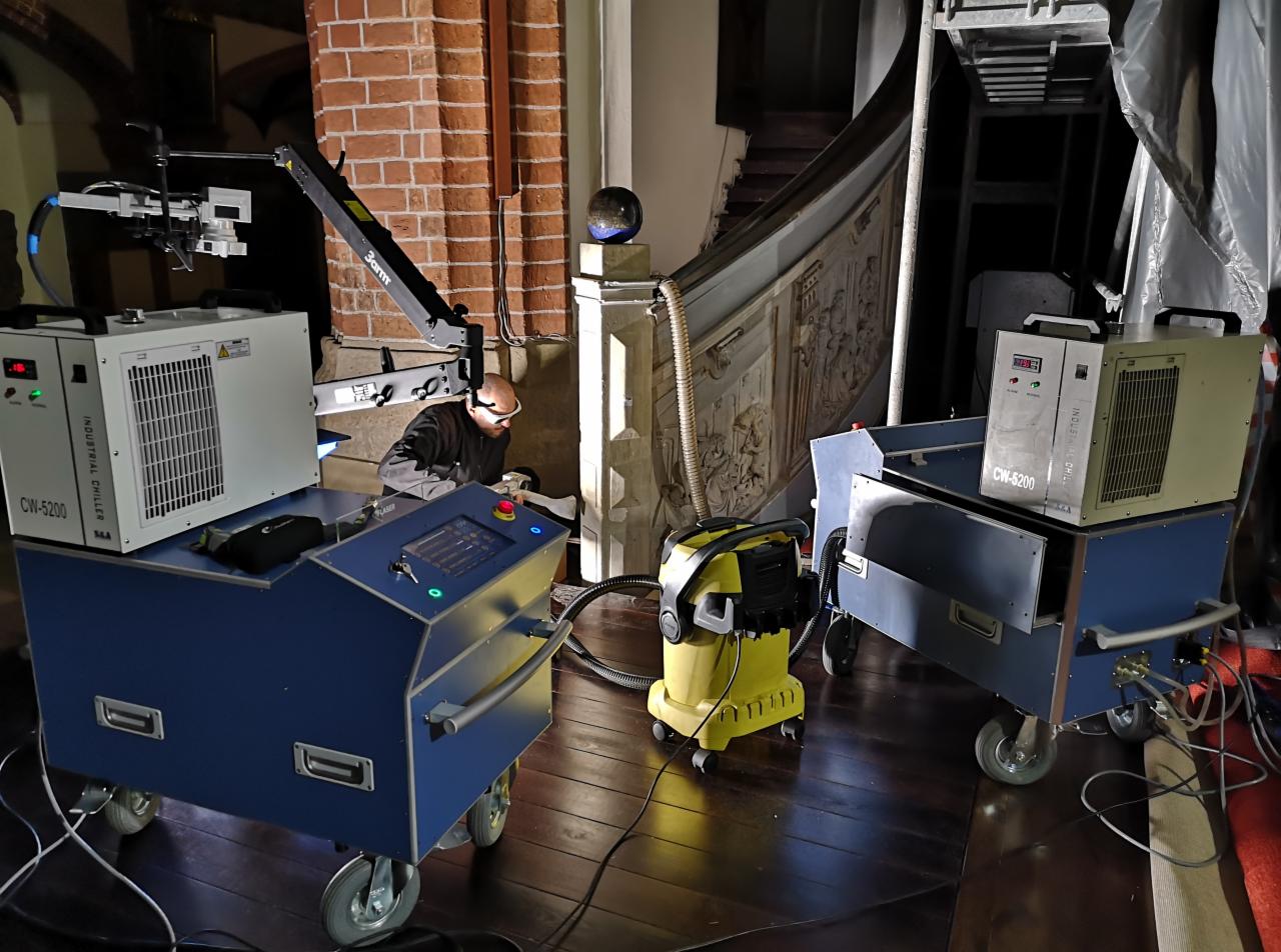 Pracownik w okularach ochronnych obsługuje laserowy system czyszczący z chłodnicą przemysłową CW-5200, podczas renowacji zabytkowych schodów z płaskorzeźbami w budynku z cegły.