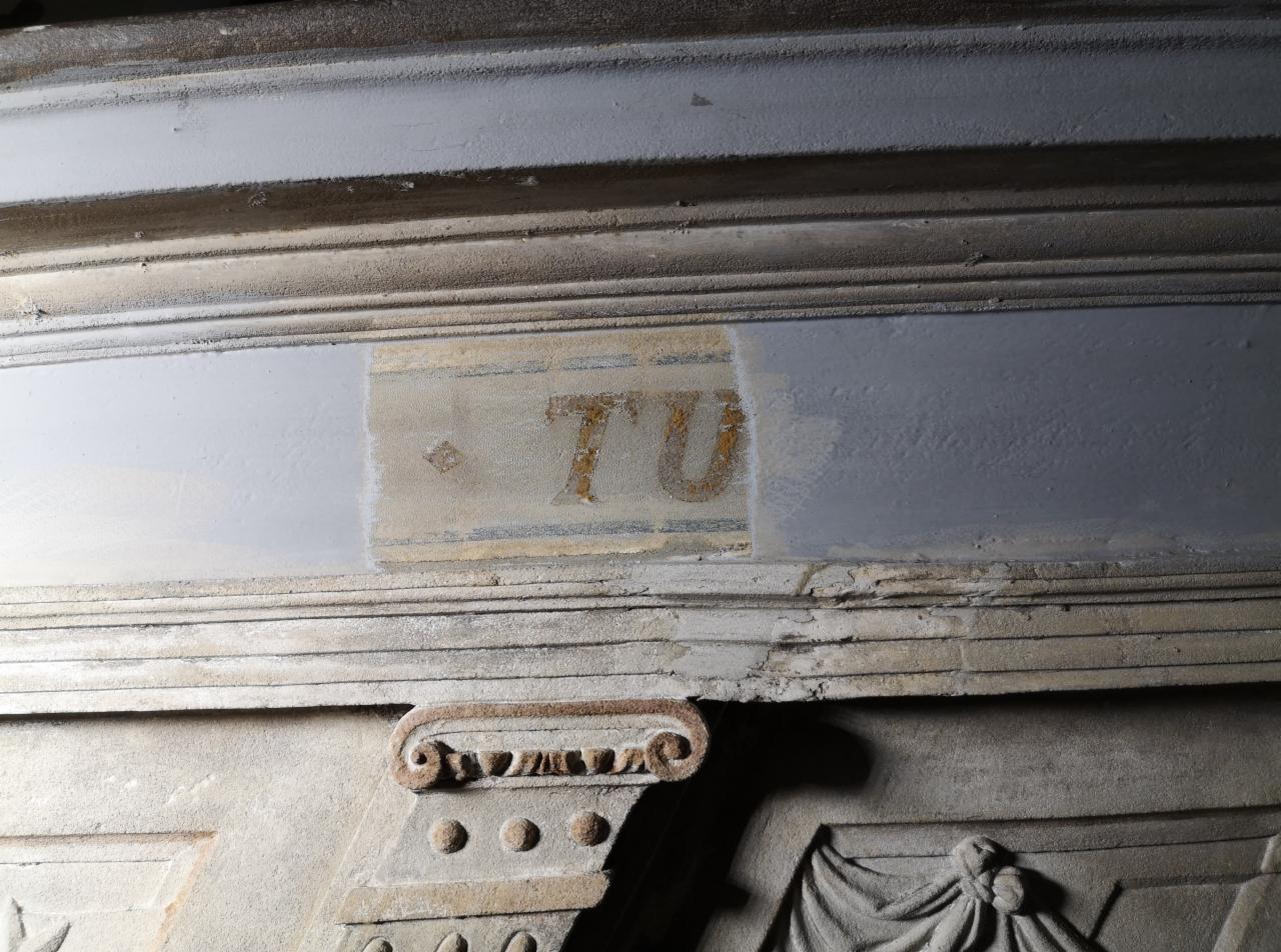 Fragment odrestaurowanego, zabytkowego elementu architektonicznego z inskrypcją 'TU' w trakcie prac konserwatorskich, widoczne ślady czyszczenia i uzupełniania ubytków.