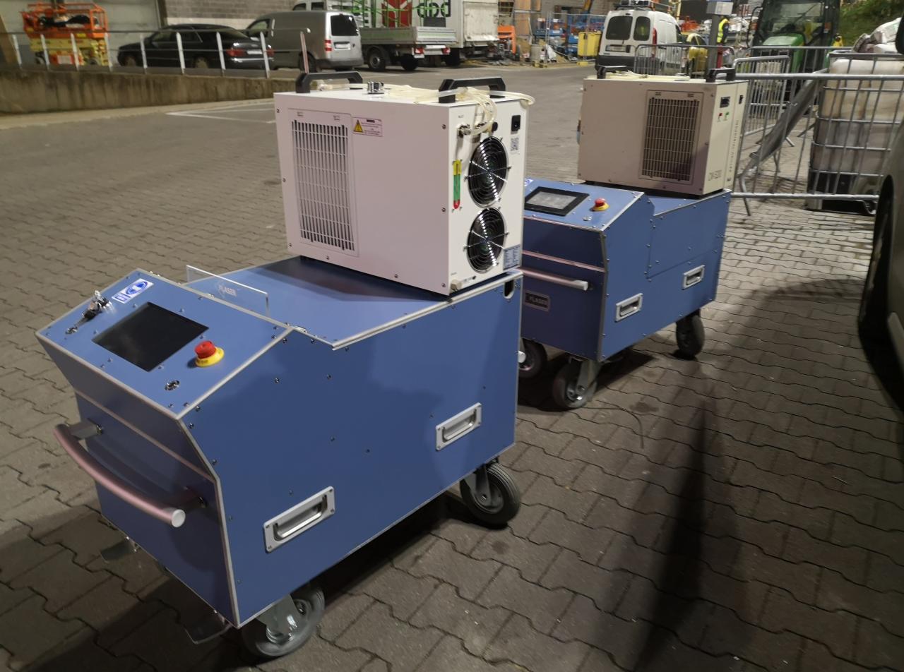 Dwa mobilne urządzenia przemysłowe w kolorze niebieskim z białymi agregatami chłodniczymi na górze, stojące na zewnątrz na kostce brukowej w otoczeniu samochodów dostawczych.