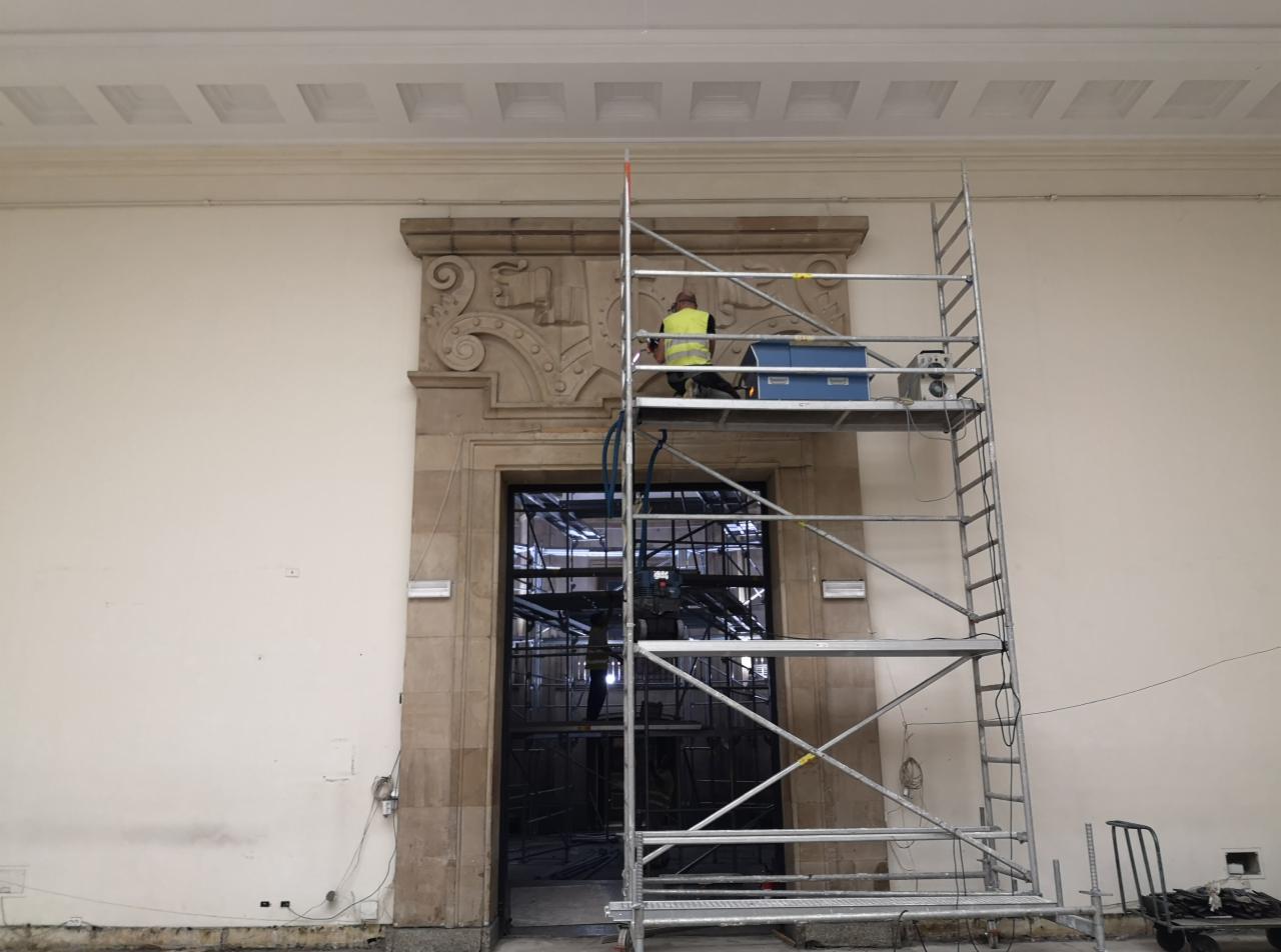 Pracownik na rusztowaniu, w żółtej kamizelce, wykonuje prace konserwatorskie przy ozdobnym portalu drzwiowym w budynku z wysokim sufitem.