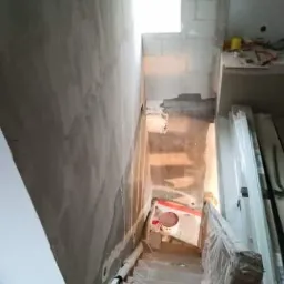 Klatka schodowa w trakcie prac przygotowawczych do wykończenia: ściany częściowo pokryte szarą gładzią, zabezpieczone schody, widoczne materiały budowlane i puszka z farbą.