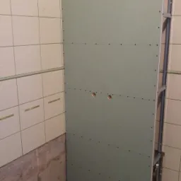 Wnętrze łazienki w trakcie prac: ściana z płyty karton-gips na metalowym stelażu, z widocznymi otworami na instalację, częściowo wykończona płytkami ceramicznymi, z odsłoniętym fragmentem ściany...