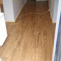 Korytarz z nową, drewnopodobną podłogą z płytek ceramicznych ułożonych w prostokątnym wzorze, ściany pomalowane na szaro, drzwi zabezpieczone folią ochronną, widoczny kabel elektryczny na podłodze.