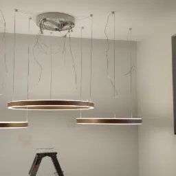 Montaż wiszących, okrągłych lamp LED z brązową obudową, widoczne kable, drabina aluminiowa i mała drabinka monterska, okno z widokiem na zieleń.