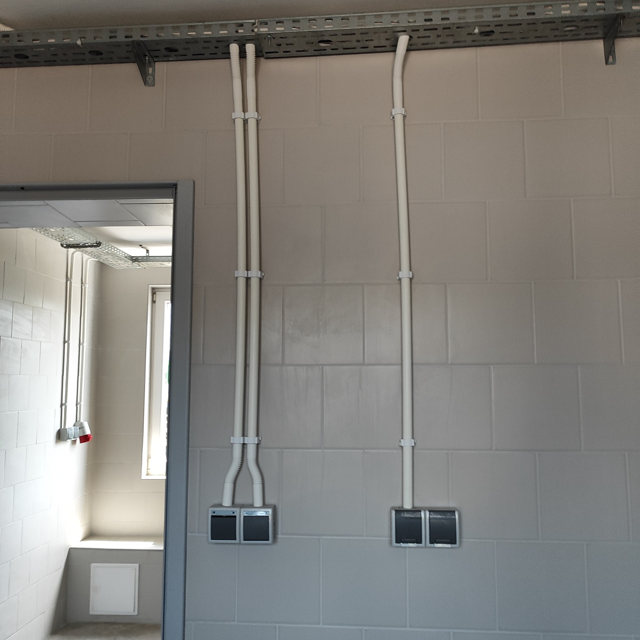 Białe rury instalacyjne prowadzone po ścianie z szarej glazury, mocowane klamrami, z metalową listwą montażową pod sufitem i widocznymi puszkami elektrycznymi podłączonymi do rur.