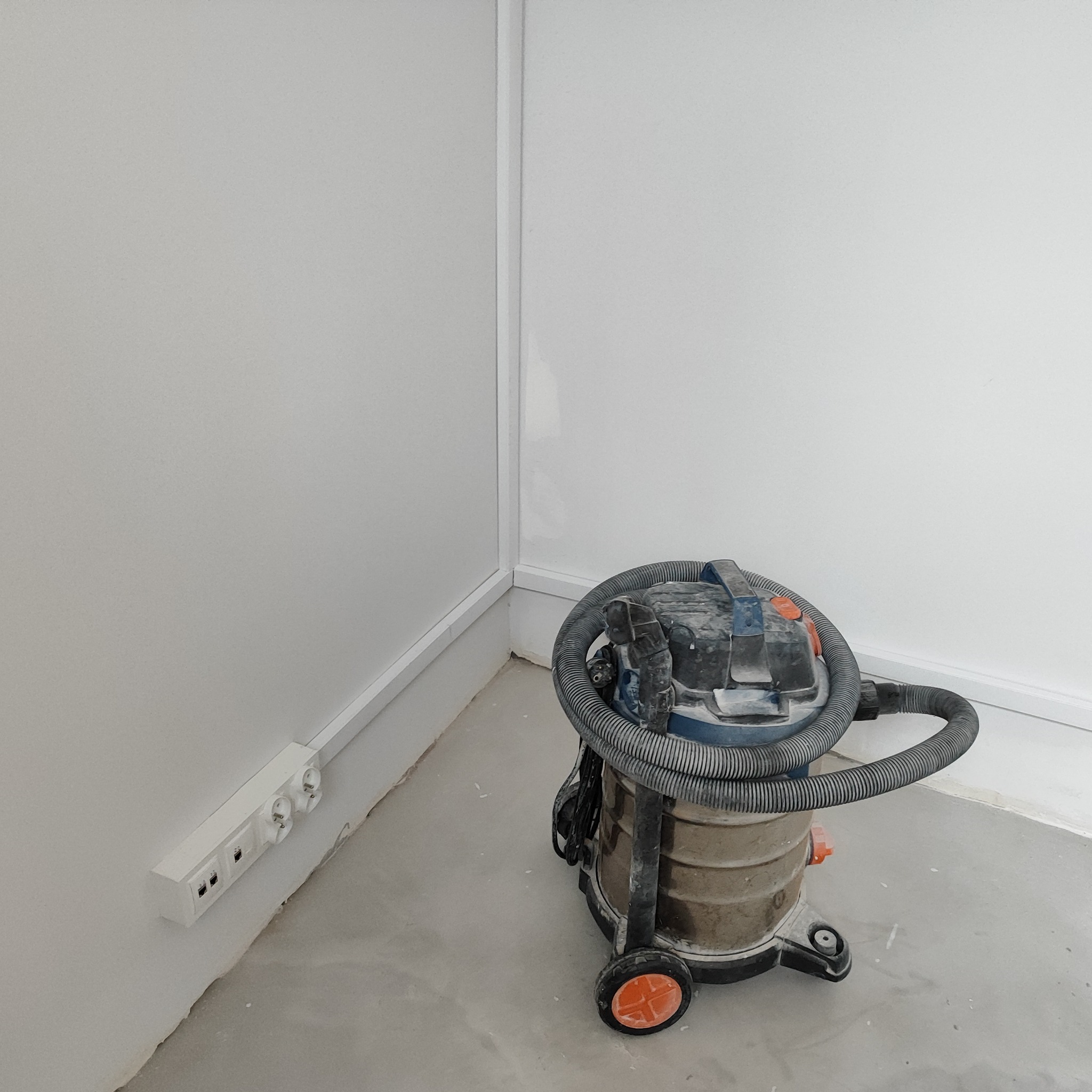 Brudny odkurzacz przemysłowy na kółkach stoi w rogu pomieszczenia z zamontowanym gniazdkiem elektrycznym i listwami przypodłogowymi.