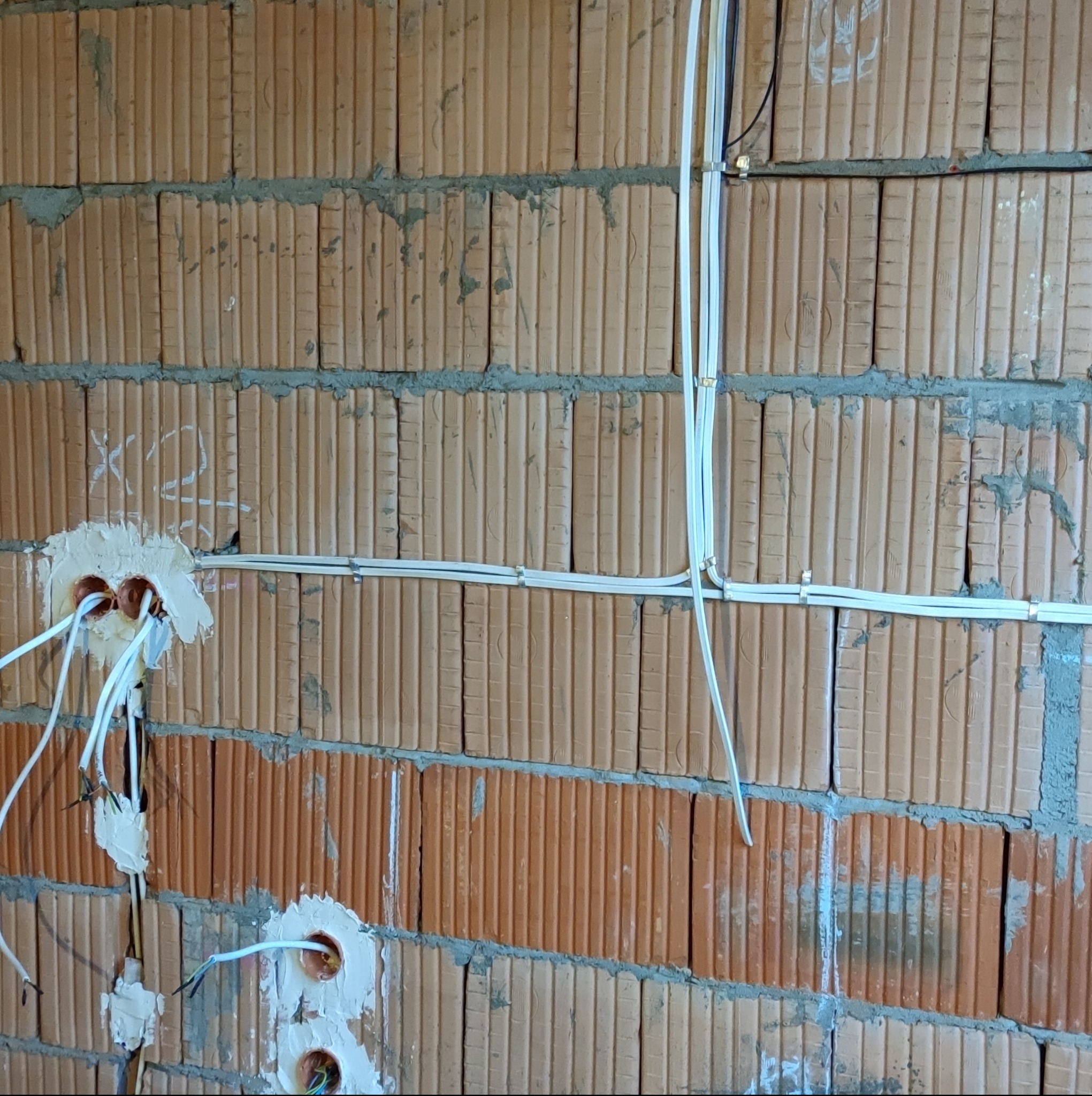 Surowa ściana z cegły z widocznymi przewodami elektrycznymi, puszki instalacyjne zamurowane z wystającymi kablami, kable przytwierdzone klamrami do ściany, etap prac instalacyjnych w budynku.