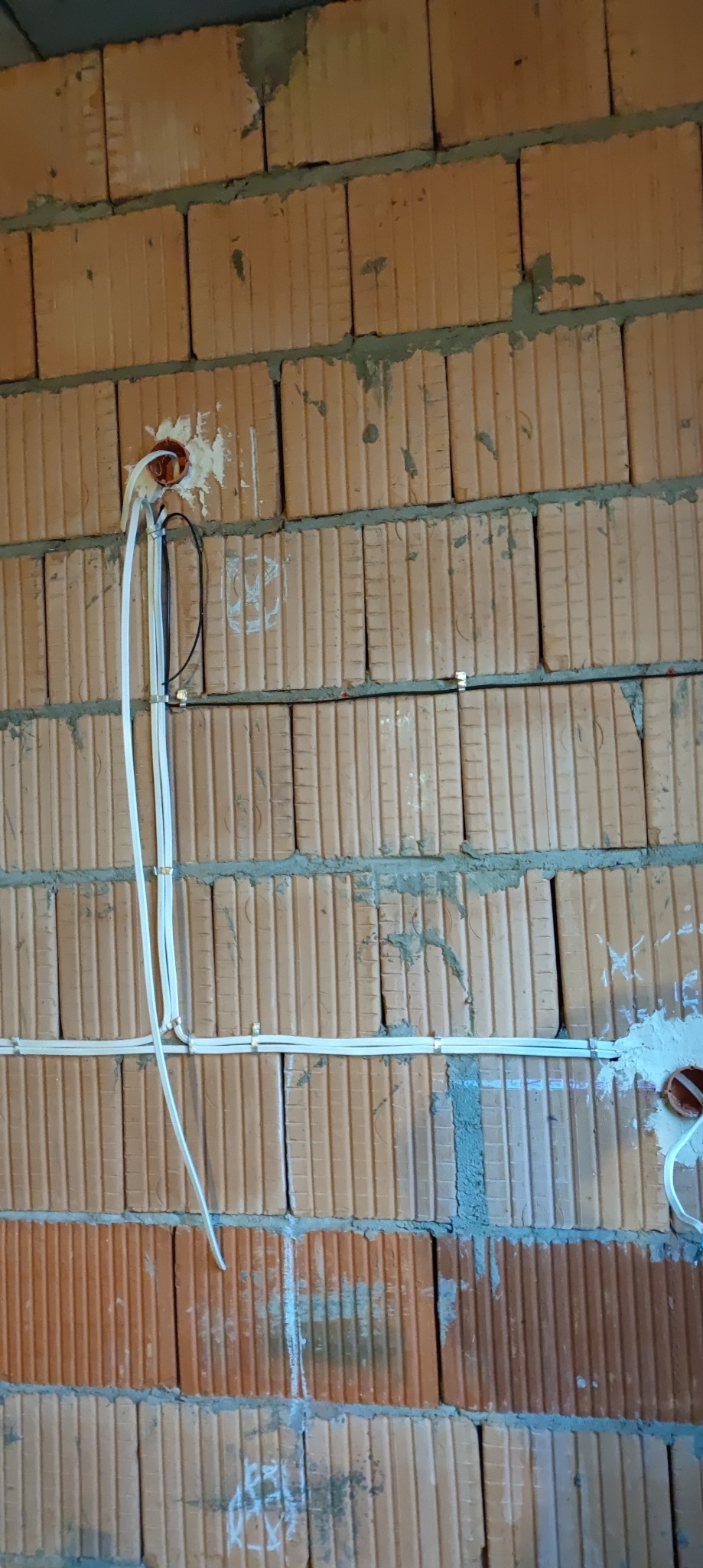 Ściana z cegły dziurawki w trakcie prac elektrycznych: widoczne przewody w peszlach przymocowane metalowymi uchwytami, puszka instalacyjna zalana gipsem.
