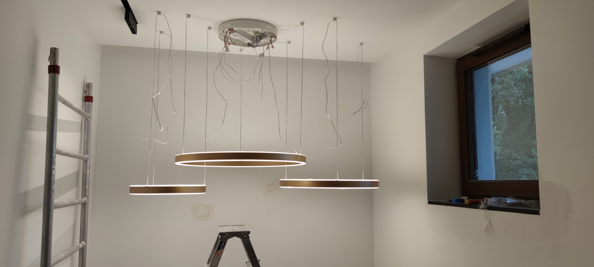 Montaż wiszących, okrągłych lamp LED z brązową obudową, widoczne kable, drabina aluminiowa i mała drabinka monterska, okno z widokiem na zieleń.