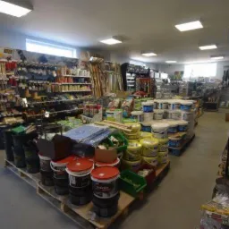 Wnętrze sklepu budowlanego z szerokim asortymentem farb, narzędzi i materiałów wykończeniowych na półkach i paletach, oświetlonych jasnym, równomiernym światłem.