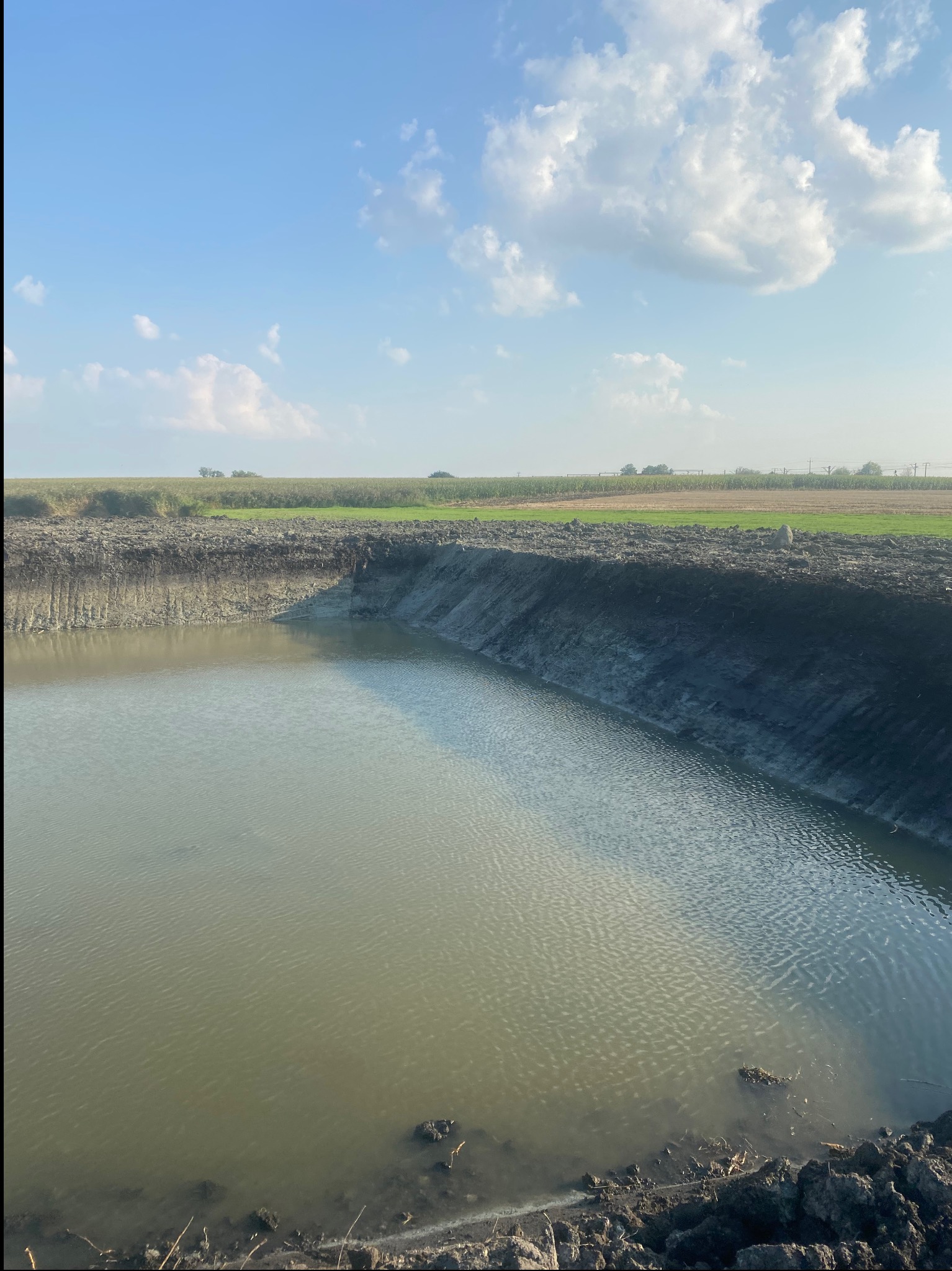 Wykopany staw wypełniony wodą, z widocznym brzegiem z ziemi i tłem z zielenią i błękitnym niebem z chmurami. Ujęcie z góry, podkreślające rozmiar wykopu.