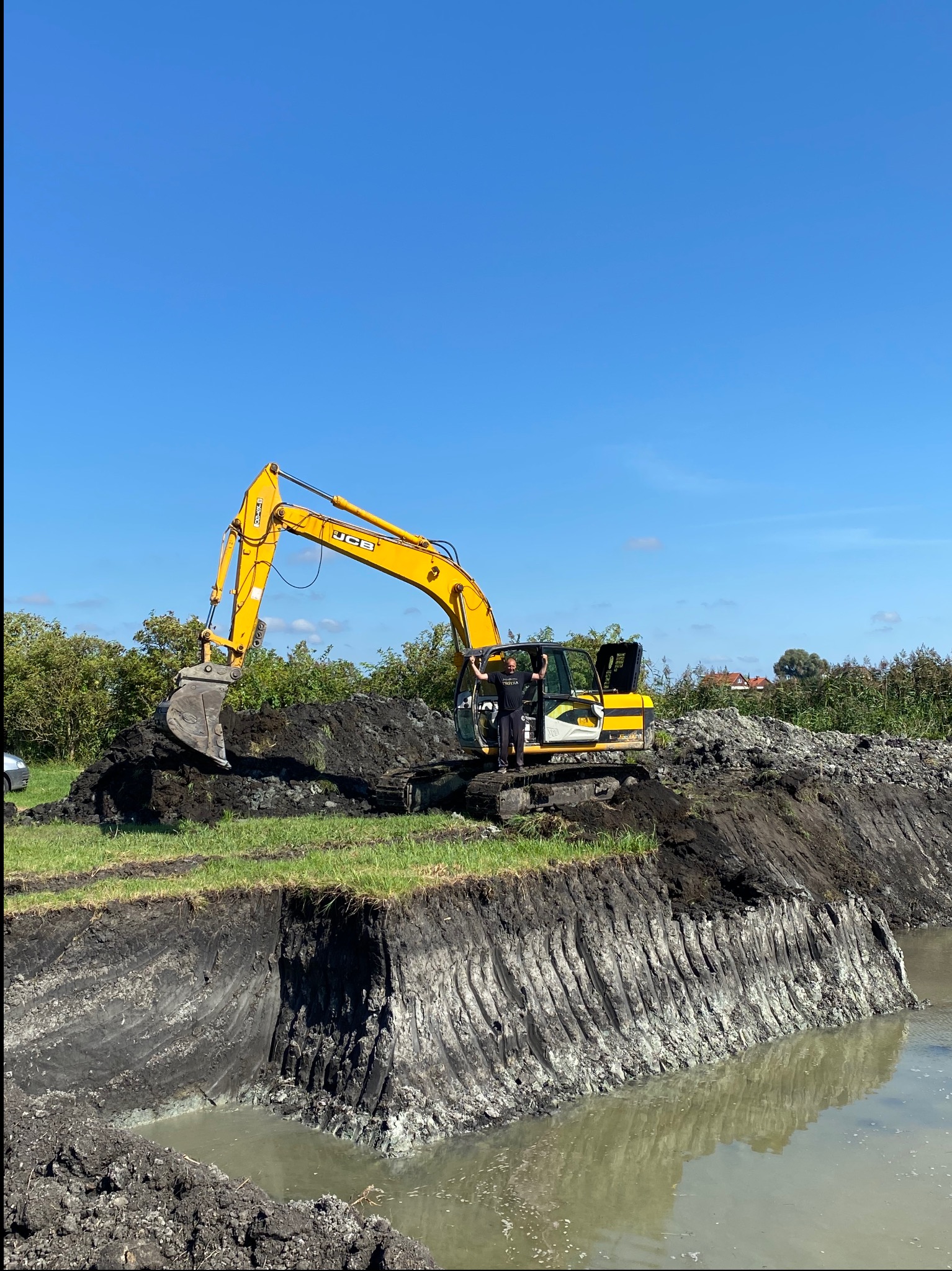 Żółta koparka JCB przy wykopanym stawie, operator stoi w kabinie z podniesionymi rękami. Widoczne warstwy ziemi i odbicie w wodzie. Błękitne niebo.