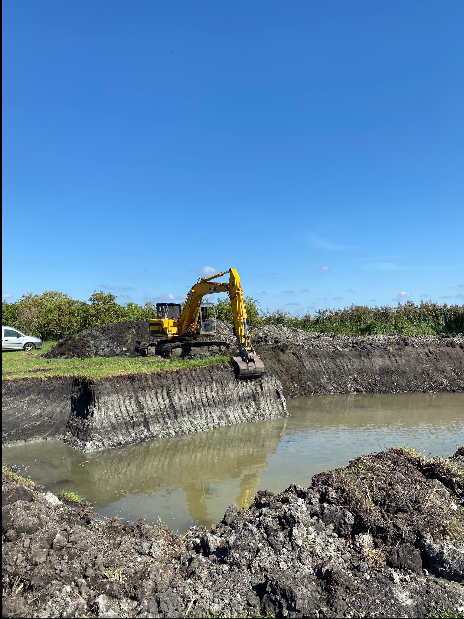 Żółta koparka JCB pogłębia częściowo wypełniony wodą staw, widoczne ślady pracy na brzegach, w tle zieleń i błękitne niebo. Prace ziemne w toku.